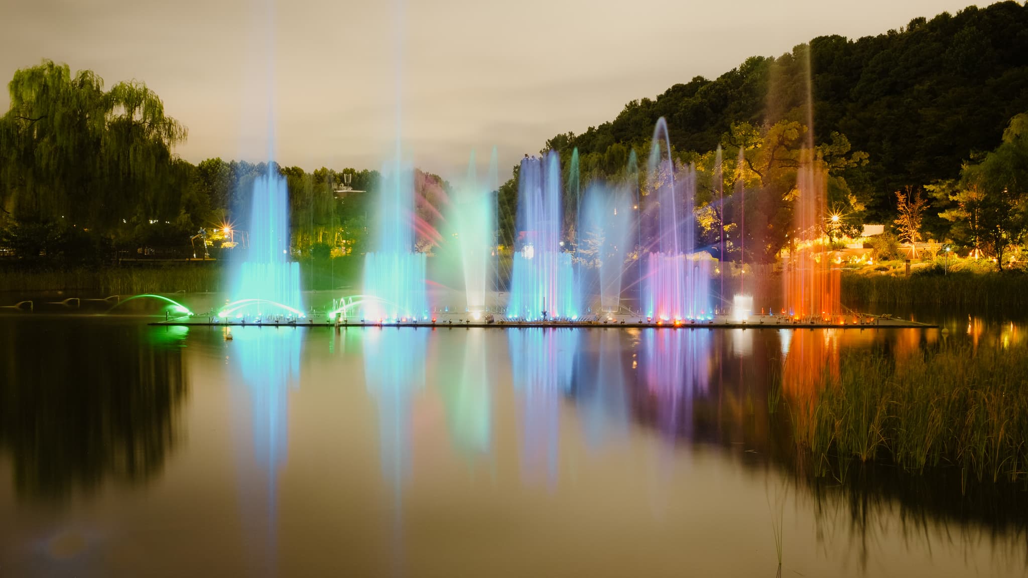 Colorful illuminated fountains reflecting on a calm lake at night, surrounded by trees.