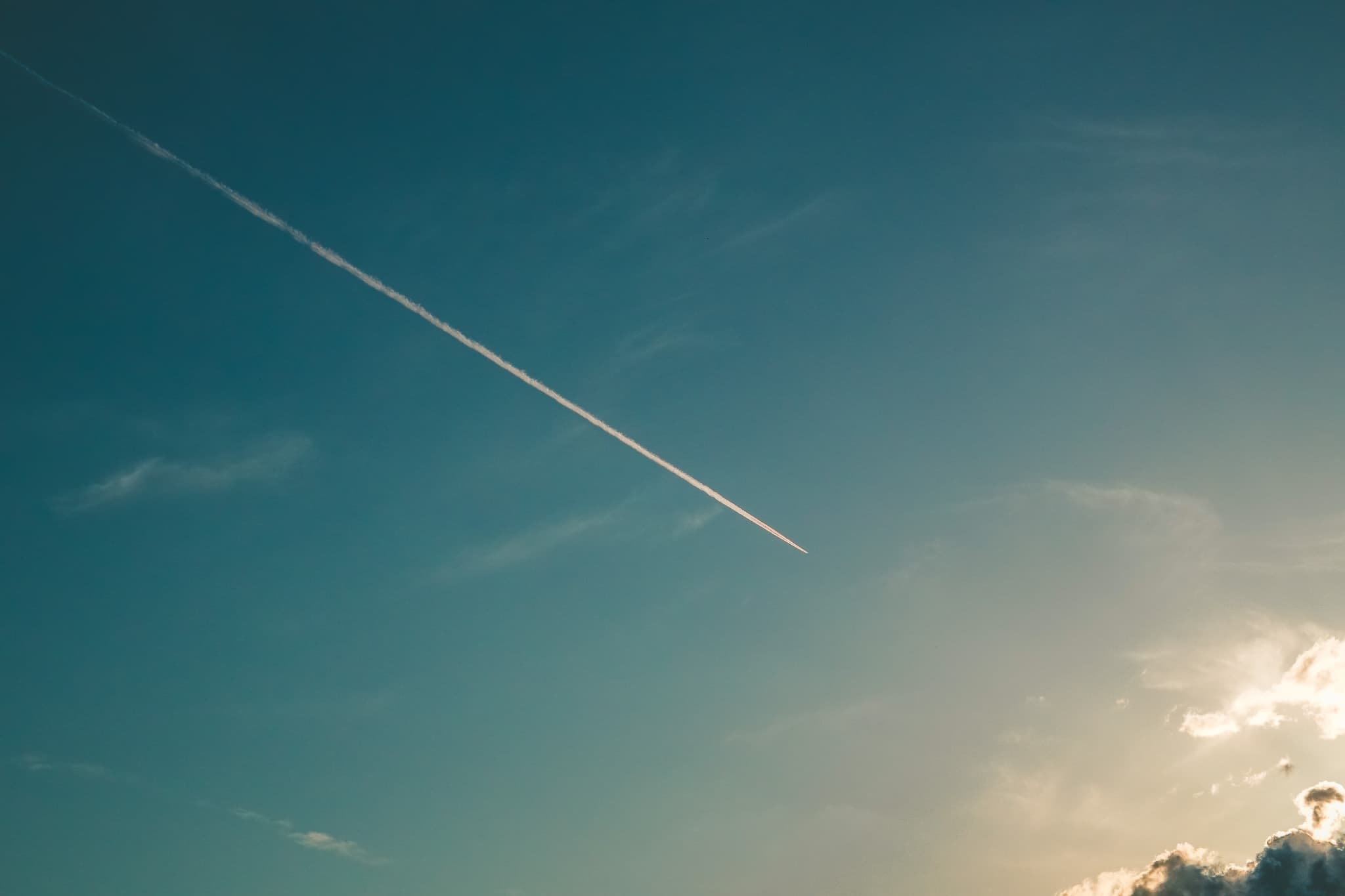 Jet contrail streaks across a clear blue sky as the sun begins to set, casting a warm glow on scattered clouds.